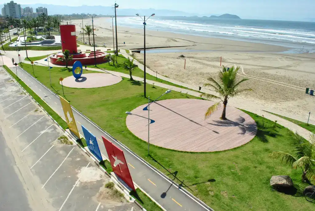 Aerial perspective of a coastal park and promenade. The area features vibrant green lawns with circular paved wooden decks, a tall red cylindrical monument, and stylized blue and yellow sculptures. A paved multi-use path runs alongside a sandy beach and the ocean, with a palm tree casting a long shadow across the grass.
