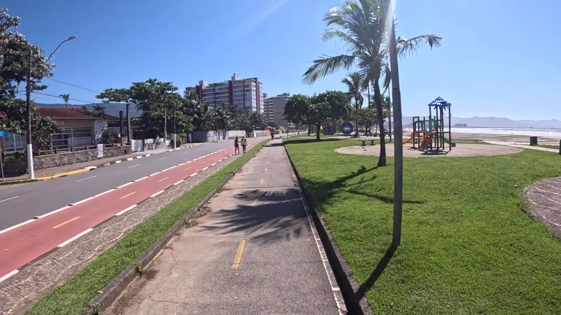 Ground-level view of a sunny beachfront promenade. A paved walking path runs parallel to a red-painted bicycle lane and a paved road. On the right side is a grassy lawn featuring a children's playground and a palm tree, leading to a sandy beach. High-rise residential buildings are visible in the background beneath a clear blue sky. Two people are walking in the distance.