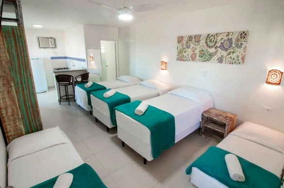 Spacious tropical dormitory room with six twin beds featuring green and white linens, a small kitchenette with bar stools, and rustic wooden accents.