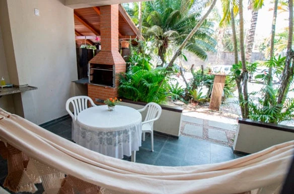 Relaxing covered outdoor patio with a white round table, chairs, a brick barbecue grill, a cozy hammock, and lush green plants surrounding the area.