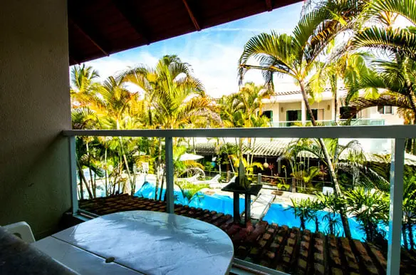 Private hotel balcony featuring a small table and chairs, offering a scenic view of the swimming pool and tropical palm tree garden below.