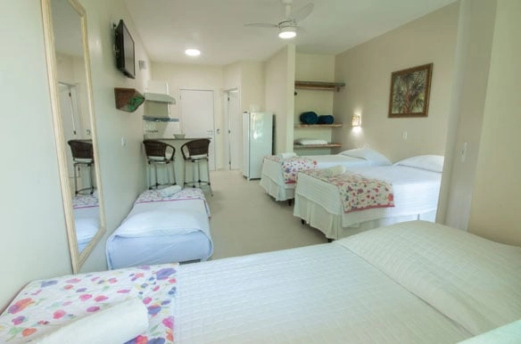 Spacious studio accommodation with four beds arranged around the room, a mirrored wall, a tall ceiling fan, and a compact kitchenette with bar stools.