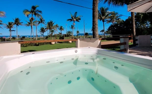 Large outdoor hot tub with bubbling water overlooking a tropical coastal landscape with palm trees, green lawns, and the ocean in the background.