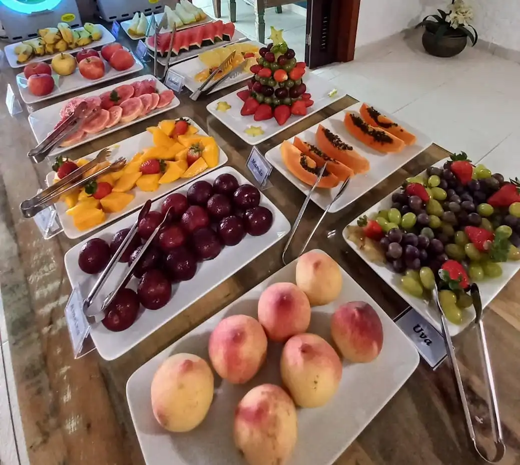 Fresh tropical breakfast buffet featuring white platters of red grapes, peaches, sliced mango, papaya, and a festive fruit tower with strawberries under bright lighting.