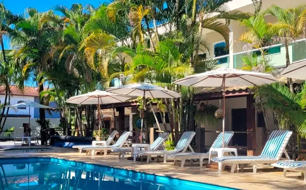 Relaxing poolside area featuring blue-and-white striped lounge chairs, large white umbrellas, and lush tropical greenery bordering the hotel building.