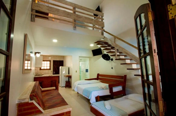 Unique multi-level loft room with white walls, a wooden staircase leading to an upper bunk area, a lower bed, and a small kitchenette corner.