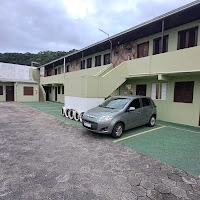 Exterior view of an apartment complex or budget pousada featuring designated paved parking spots with a parked silver hatchback and an exterior staircase.