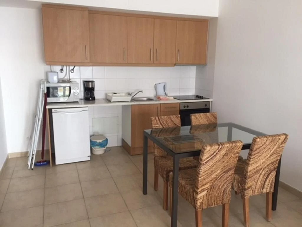 Small apartment kitchenette featuring upper wood cabinets, a white mini-fridge, microwave, coffee maker, and a glass dining table with patterned chairs.