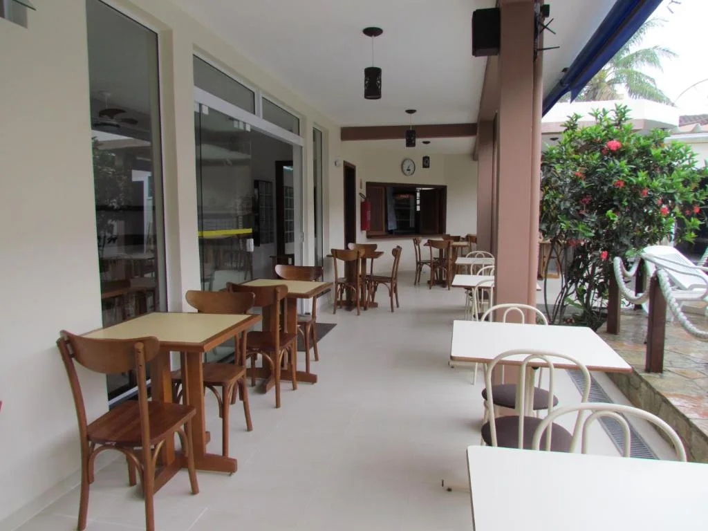 Interior view of a long, covered patio dining area with light tiled floors, wooden and white tables and chairs, sliding glass doors on the left, and lush greenery with a blue awning on the right.