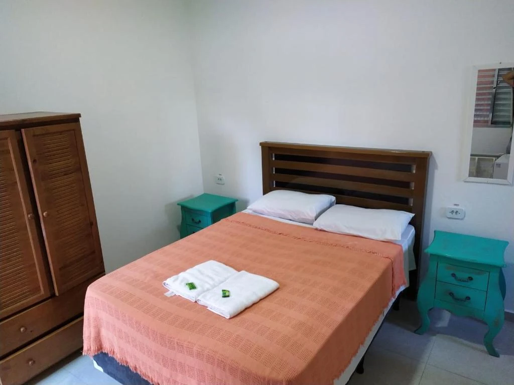 Interior view of a simple bedroom with white walls, a wooden slatted headboard, an orange fringed bedspread, white towels on the bed, a wooden wardrobe, and teal nightstands.