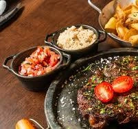 Close-up of a sizzling black skillet steak with tomatoes, served with farofa, salsa, and fries.