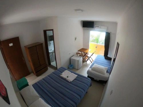 Wide-angle interior view of a compact bedroom featuring a bed with a blue striped bedspread, a single bed, a wooden wardrobe, a mini-fridge, a wall-mounted TV, and an open balcony door.