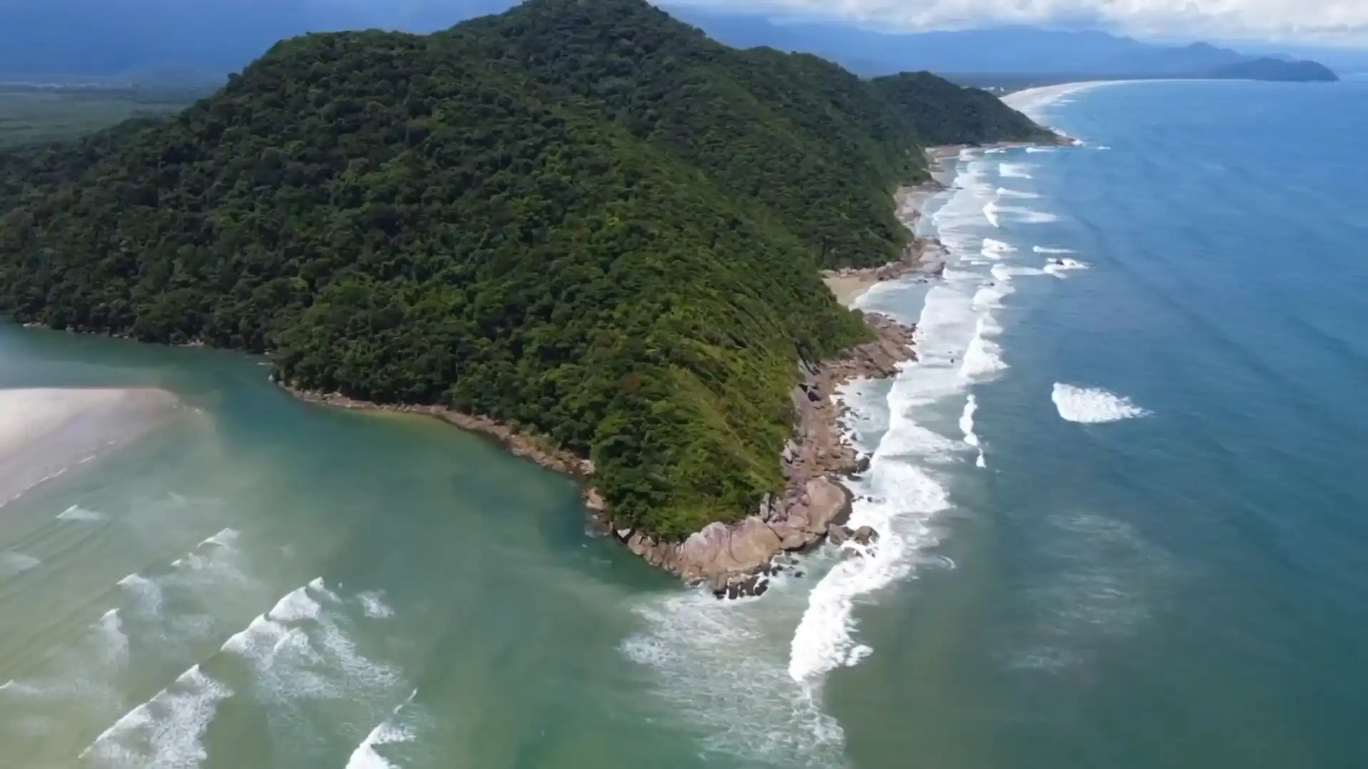 Aerial drone view of a dense green mountain sloping down to a rocky coastline with white crashing waves and blue ocean water.