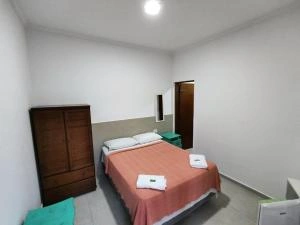 Wide-angle interior view of a minimalist bedroom featuring a bed with peach-colored bedding, white towels, a dark wooden wardrobe, a mirror, and a white ceiling light.