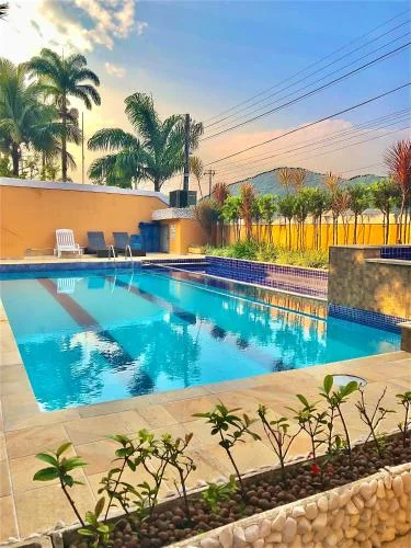 Scenic view of a clear blue swimming pool reflecting the sky, bordered by an orange wall, potted plants, palm trees, and a sunset sky with clouds.