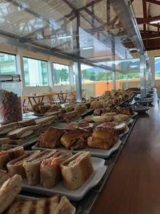 Interior view of a bright breakfast room with large glass windows, featuring a long buffet counter loaded with various breads, pastries, and cakes.