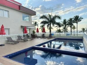 Coastal hotel pool area featuring blue mosaic pools, red umbrellas, lounge chairs, tall palm trees, and a direct view of the sandy beach and ocean.
