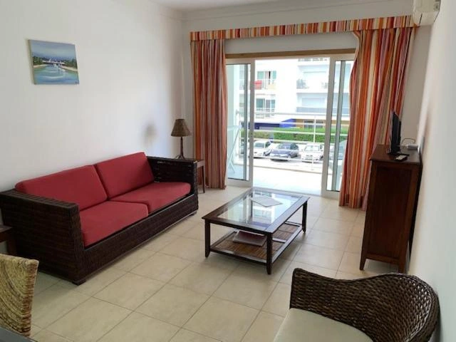 Living room interior with a red fabric sofa, glass coffee table, wooden TV stand, and sliding glass doors leading to a balcony with striped curtains.