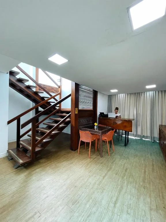 Wide-angle interior view of a spacious lobby with light wood flooring, a dark wooden staircase leading to an upper level, orange chairs around a table, and a reception desk with a staff member in the background.