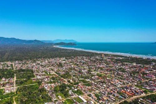 Bird's-eye perspective of a paved main road running parallel to a wide sandy beach and dense forest, with a prominent green mountain peak in the background.