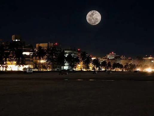 A bright, full moon shining over a dark beach shoreline with silhouetted palm trees and illuminated city buildings in the background.