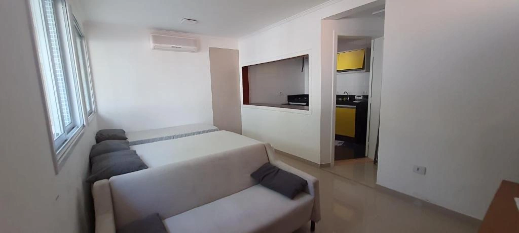 Bright studio apartment living room featuring a white sofa with grey pillows, AC unit, and a pass-through view of the yellow kitchen.