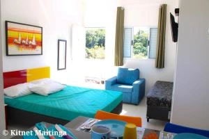 Vibrant guest bedroom featuring a teal bed with a red and yellow headboard, colorful framed art, a blue armchair, and a window with green nature views.