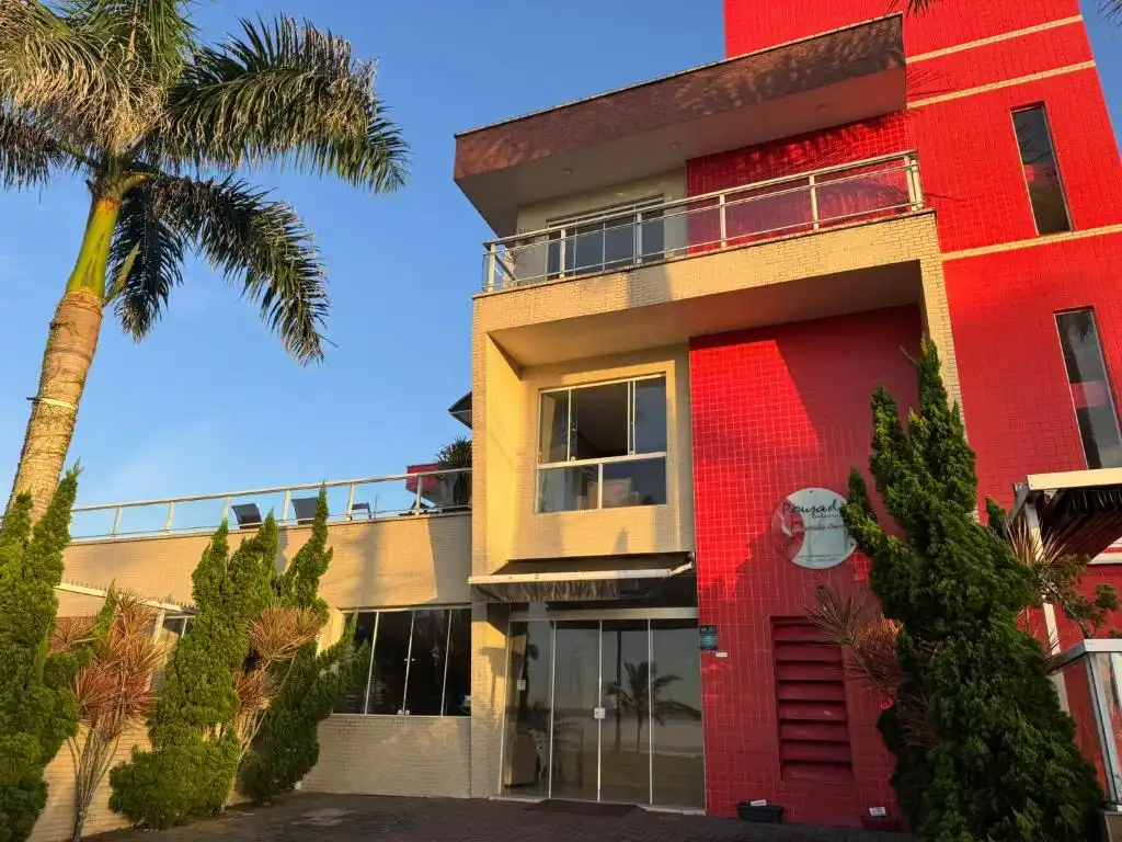 Contemporary hotel exterior with a vibrant red tiled section, beige brick structure, glass balcony, and mature palm trees catching golden hour light.