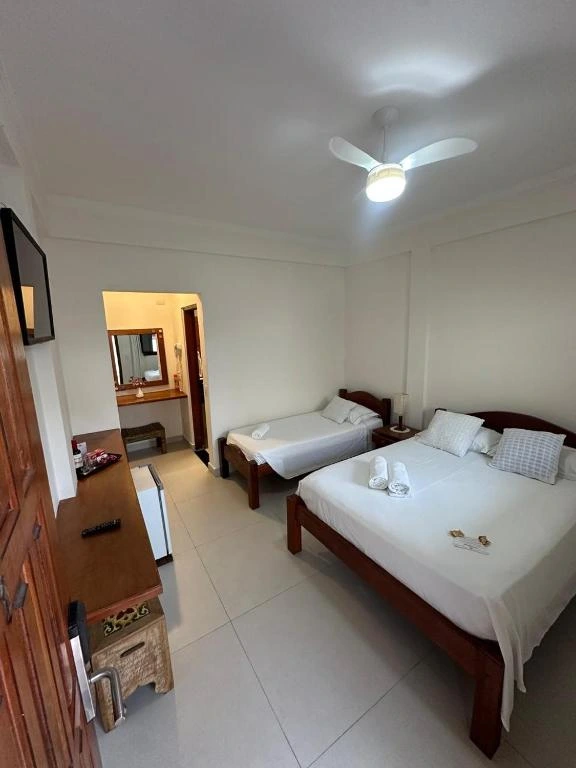 Interior of a bright bedroom with two single beds, white bedding, a wall-mounted TV, and a wooden dresser with a mini-fridge, leading to an ensuite vanity area.