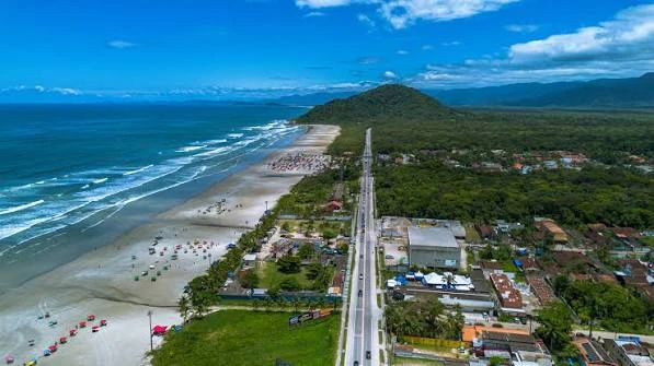High-angle aerial shot of a sprawling beachfront town bordering a vast, deep green forest and a sweeping coastline with rolling ocean waves.