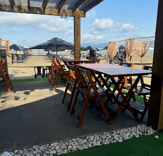 Wooden outdoor tables and chairs under a pergola, overlooking a sandy beach with cabanas and clear blue sky.