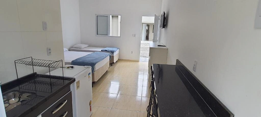 Small studio layout showing a black countertop kitchenette, white mini-fridge, and two twin beds with blue blankets and a wall-mounted TV.
