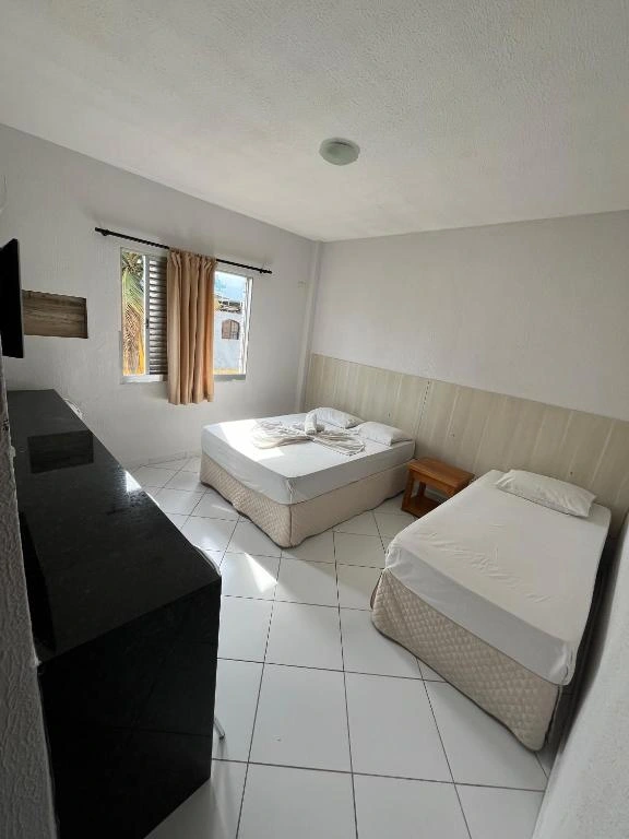 Clean white hotel room interior with two single beds, neutral bedding, wall-mounted TV, and a window with peach curtains.