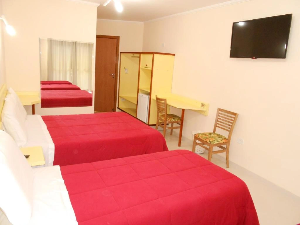 Bright hotel room featuring two red beds, a large wall mirror, a yellow wardrobe with a mini-fridge, and a wall-mounted TV.