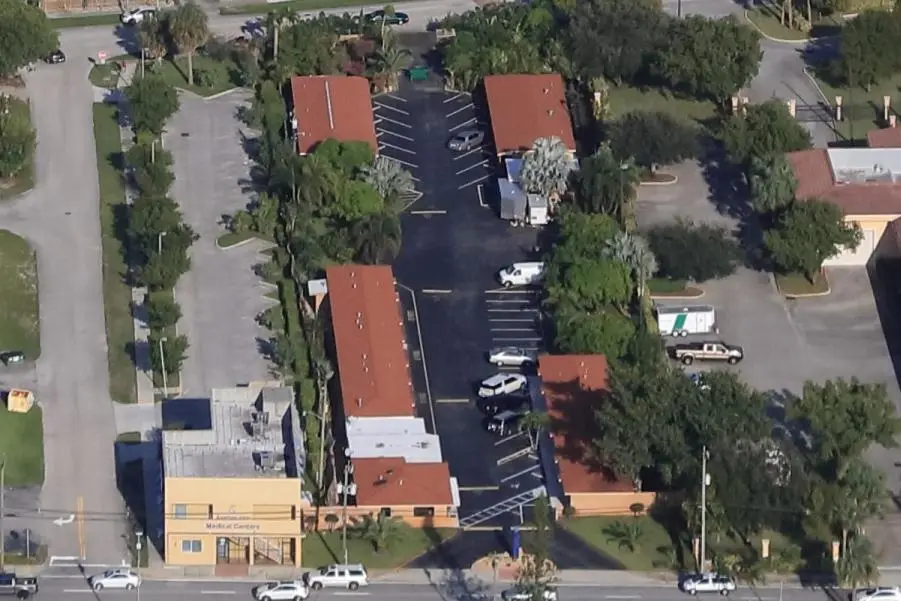 Aerial drone view of a medical building complex with red tile roofs, surrounded by trees and a large asphalt parking lot with cars.