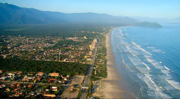 Wide aerial view of a long, empty white sand beach bordered by a paved coastal highway, dense vegetation, and calm blue ocean waters.
