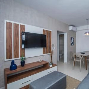 Open-plan living space showing a wood-paneled TV wall with a mounted television, a grey console table, a grey ottoman, and a dining area in the background.