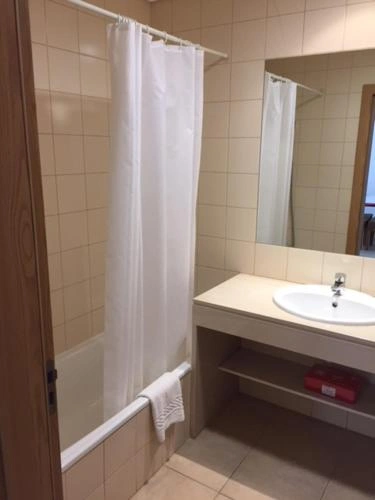 Functional bathroom featuring beige wall tiles, a white bathtub with a shower curtain, and a white vanity sink with an open shelf beneath it.