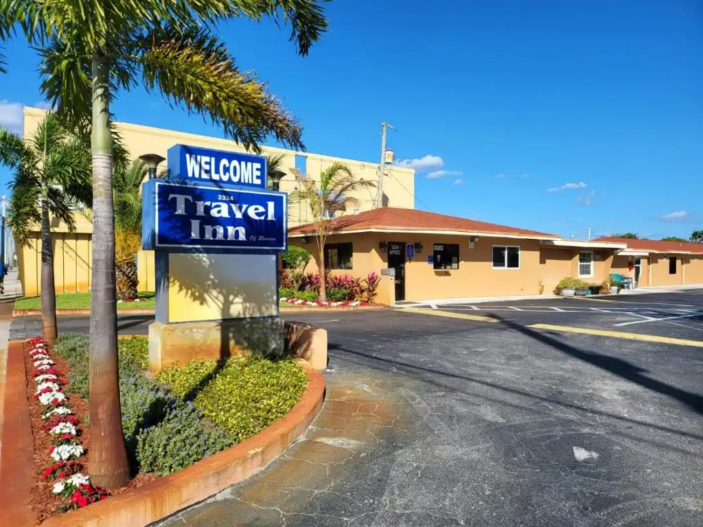 The "Welcome Travel Inn" blue and white sign standing by the entrance with palm trees and landscaping under a clear blue sky.