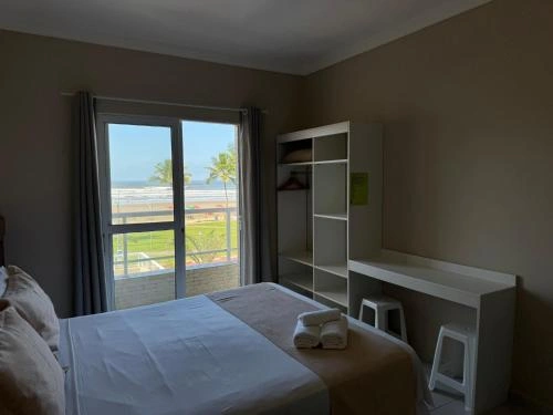 Bright, airy hotel room with a comfortable bed, rolled white towels, open closet shelving, a built-in desk, and floor-to-ceiling sliding glass doors facing the beach.