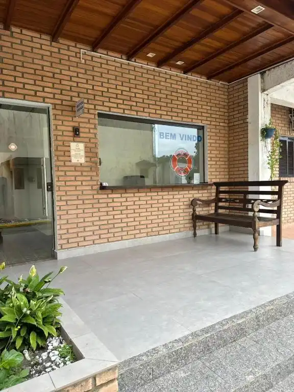Exterior view of a lodge reception area featuring a brick wall, a glass window with a 'Bem-Vindo' sign and life ring, a wooden bench, and glass sliding doors.
