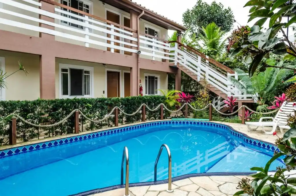 View of a two-story building with pink walls and white balconies, featuring a curved, blue-tiled swimming pool with metal steps in the foreground, surrounded by tropical plants and a rope fence.