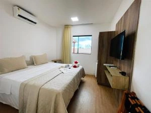 Interior view of a bright, air-conditioned bedroom with a double bed, light tan bedding, white pillows, a wooden dresser with a wall-mounted TV, and a window with yellow curtains.