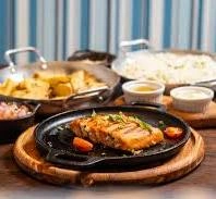 Top-down view of a sizzling salmon fillet on a hot cast-iron plate, served with rice and fried sides.