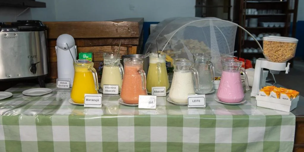 Pousada La Naila breakfast buffet display with a green gingham tablecloth, fresh fruit juices, a coffee urn, and cereal dispenser.