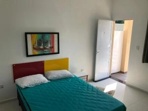 Simple, sunlit bedroom showing a bed with a teal bedspread, a unique red-and-yellow headboard, a sailboat painting, and a white door opening to the outside.