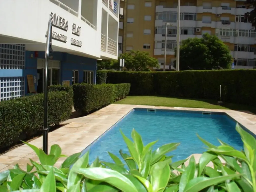 Outdoor swimming pool surrounded by beige tiled decking, lush green hedges, and the blue and white Riviera Flat Apartments building in the background.