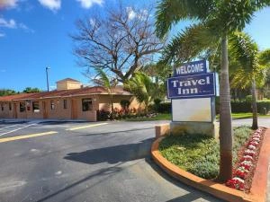 Exterior view of the Travel Inn entrance featuring the welcome sign, curved driveway, lush landscaping, and the hotel building in the background.
