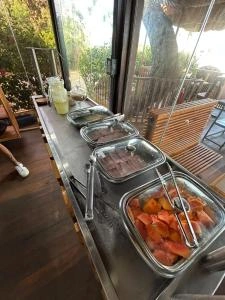 High-angle view of a breakfast buffet counter featuring stainless steel chafing dishes with hot food, tongs, and a glass of juice, set near an open window with garden views.