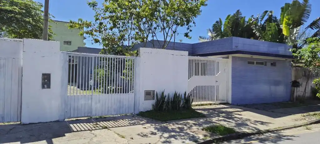 Exterior street view of a modern blue and white single-story house with a white metal gate, fence, and surrounding lush green trees.